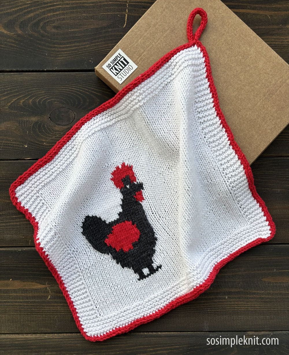 A knitted cotton dish towel with red edging and a hanging loop, featuring a black and red chicken in a jacquard pattern