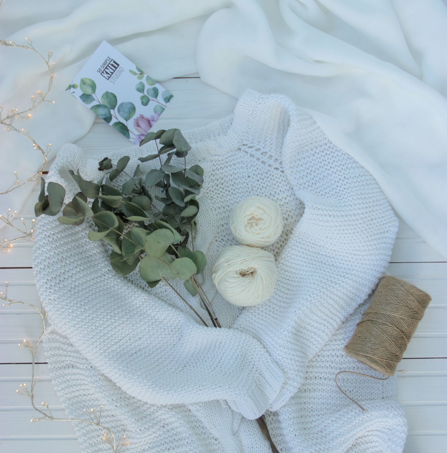 A white knitted sweater made from white yarn, beautifully arranged on a white background, with a bouquet of green eucalyptus leaves placed on top