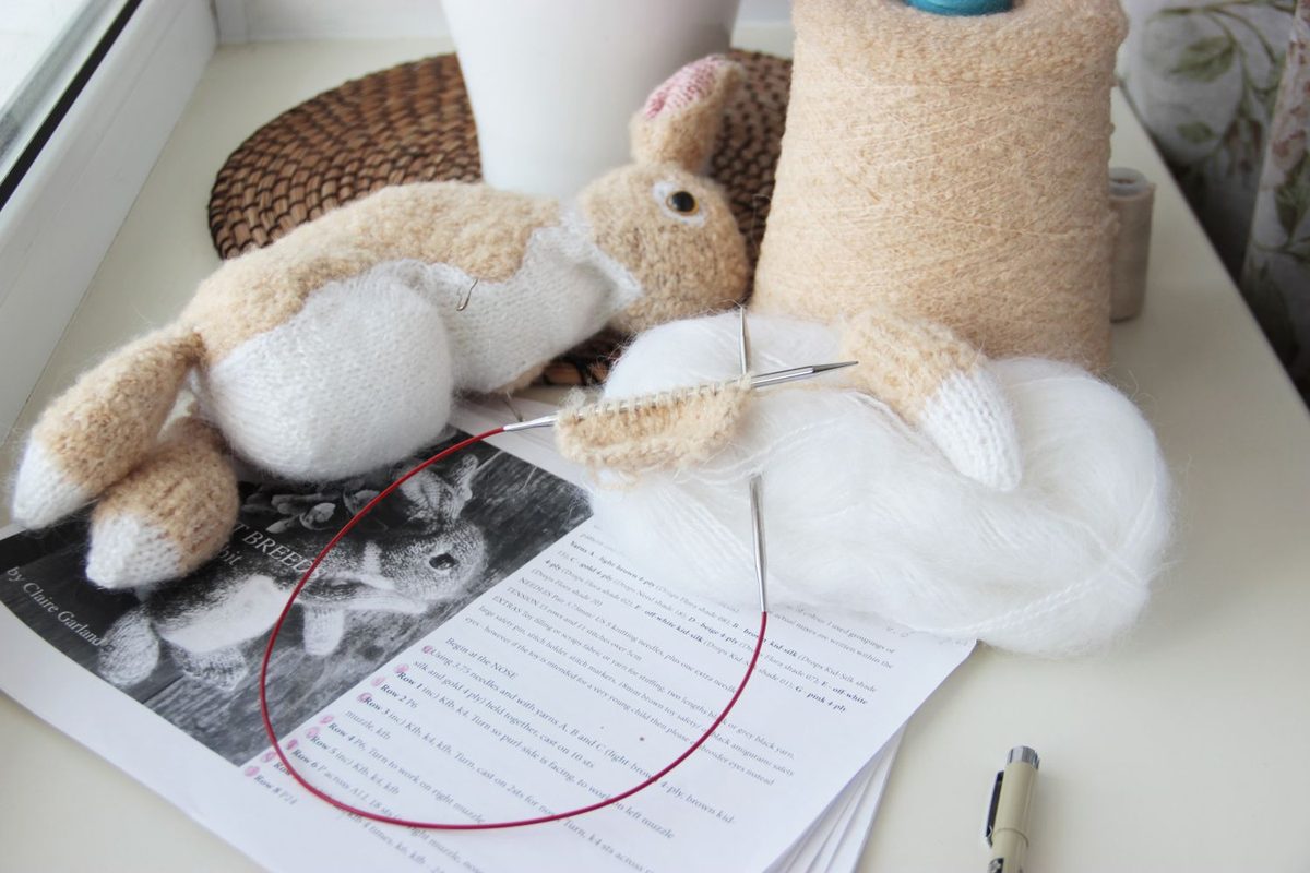 A half-knitted bunny made with needles from fluffy beige and white yarn, lying on a table, with spools of beige and white yarn nearby