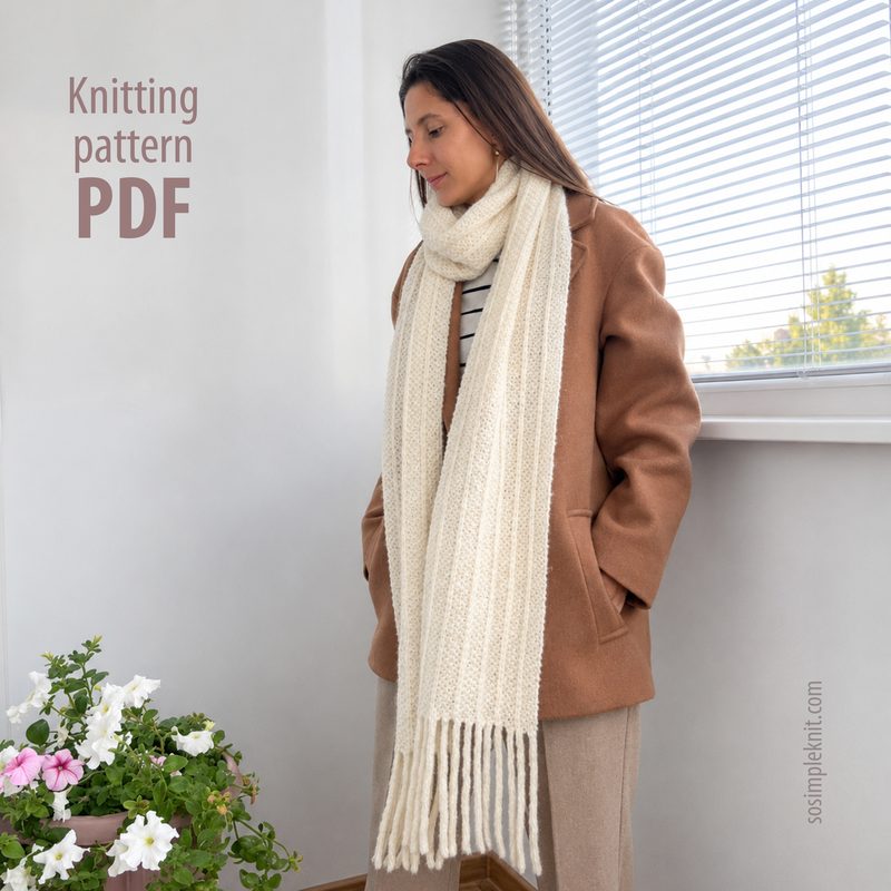 A woman stands indoors, wearing a light beige long reversible scarf draped around her neck. She is dressed in a brown oversized jacket over a striped top, with her hair cascading down. The background features a light-colored wall and a window with sheer white curtains, allowing natural light to fill the space. A potted plant with pink and white flowers is positioned to the left. The text "Knitting pattern PDF" is visible in a simple font on the right.