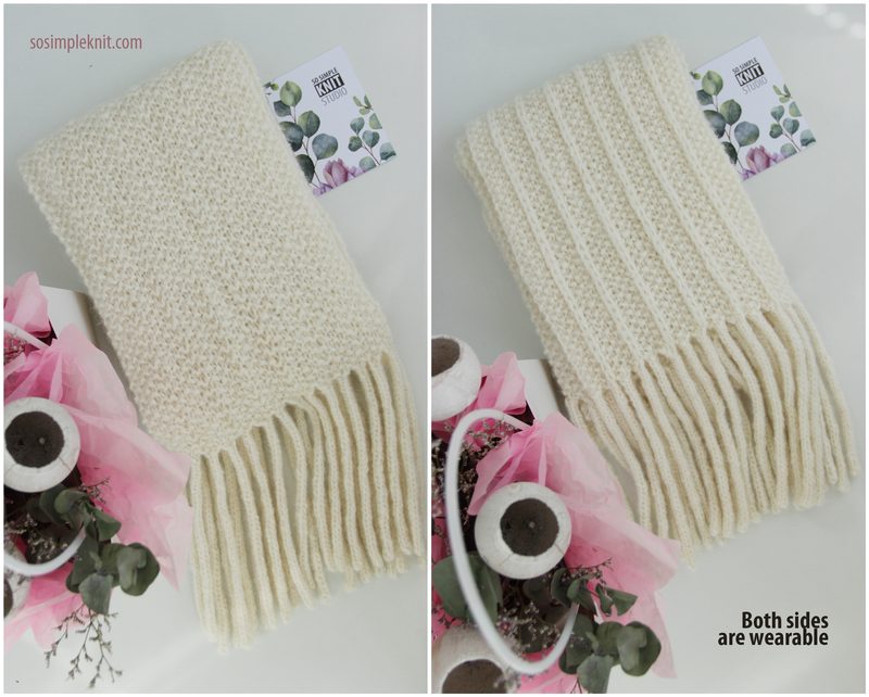 The image features a long, reversible cream-colored scarf displayed flat, with one side showcasing a textured stitch pattern and the other side featuring a ribbed design. The scarf has fringe at the ends and is accompanied by a small card with floral designs. The background is neutral, with soft pink accents from surrounding decorative elements. The scarf is neatly arranged, emphasizing its dual style.