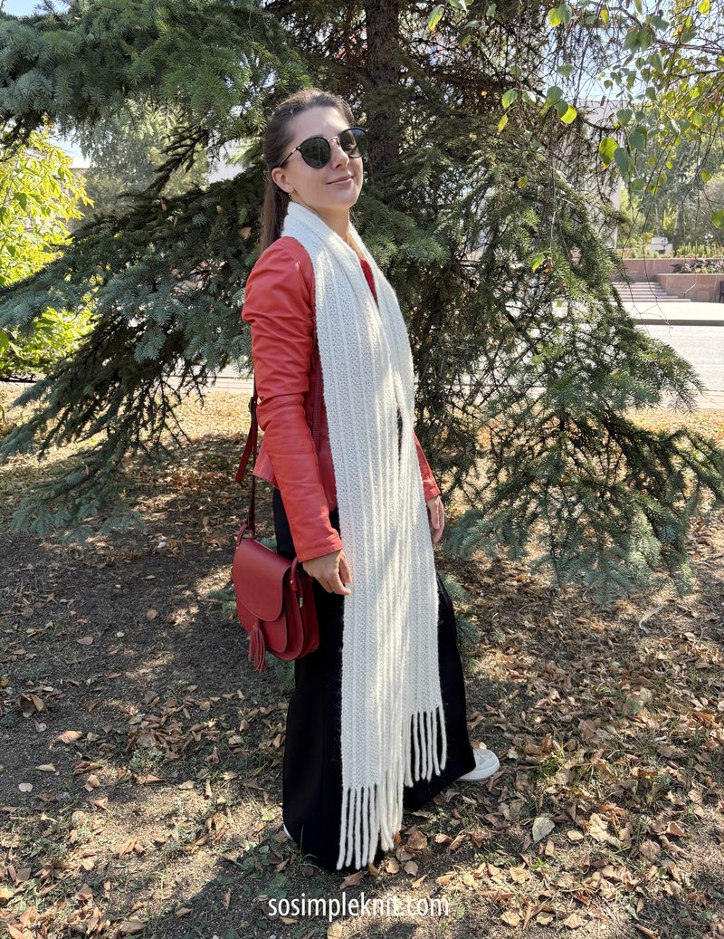 A woman stands outdoors beside a green pine tree, wearing a red leather jacket, black pants, and sunglasses. She is smiling and holding a red handbag. The long, reversible scarf she is wearing is white, featuring fringes at the ends. The background includes grass and a pathway, with sunlight filtering through the leaves.