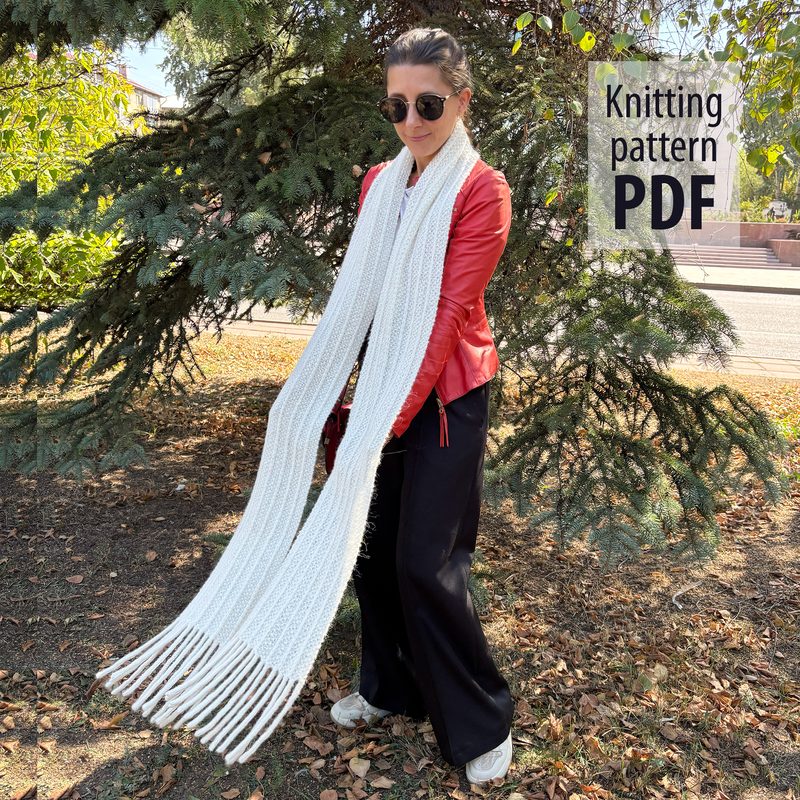 A woman stands outdoors, wearing a red leather jacket and black pants, holding a long, white reversible scarf. The scarf features a textured knit pattern and has fringe at both ends. She is framed by greenery and a glimpse of a sidewalk in the background. The text "Knitting pattern PDF" is displayed in bold black font in the upper right corner.