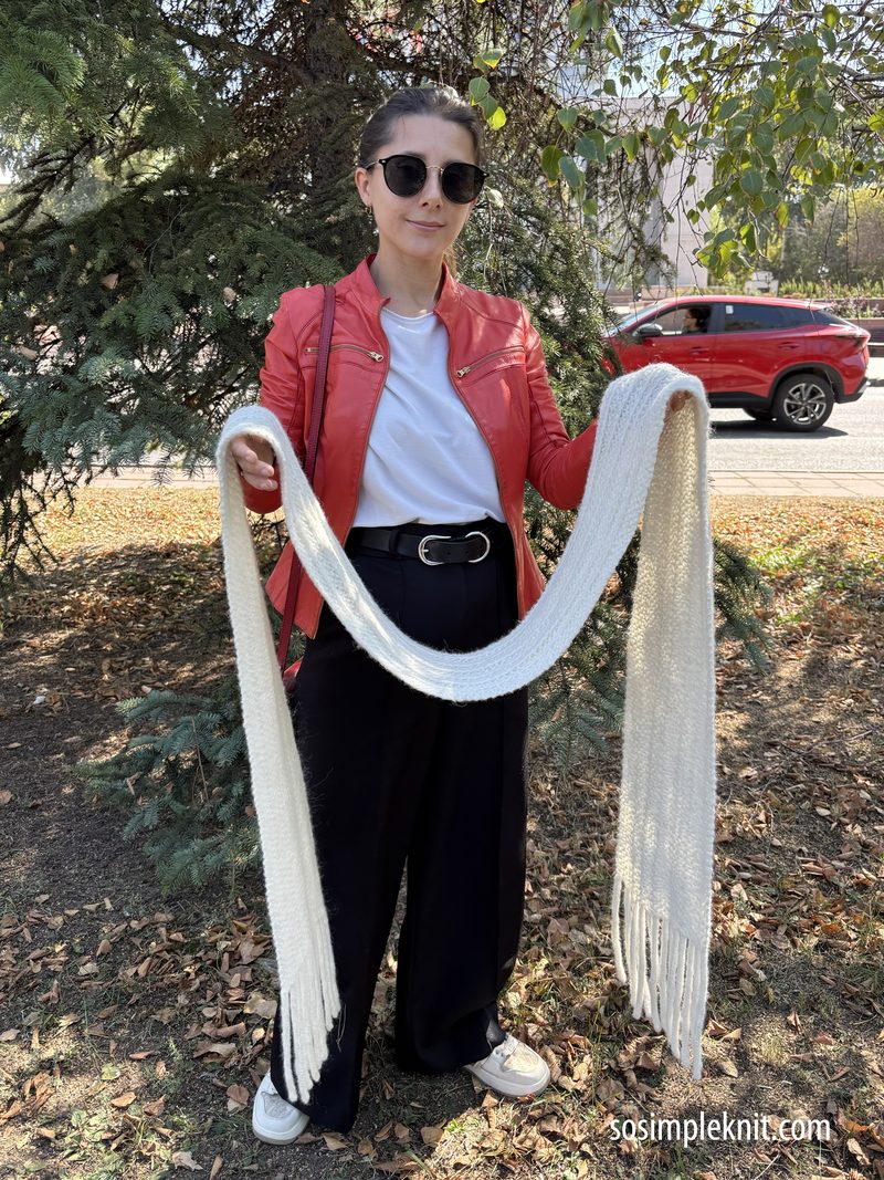 A woman stands outdoors, holding a long, white reversible scarf with fringes. She wears a red leather jacket over a white shirt, paired with black, loose-fitting pants and white sneakers. Her sunglasses are dark and stylish, and her hair is tied back. The background includes a green tree and a red car, with fallen leaves on the ground, suggesting an autumn setting. The scarf drapes in an arc between her hands, showcasing its length and texture.