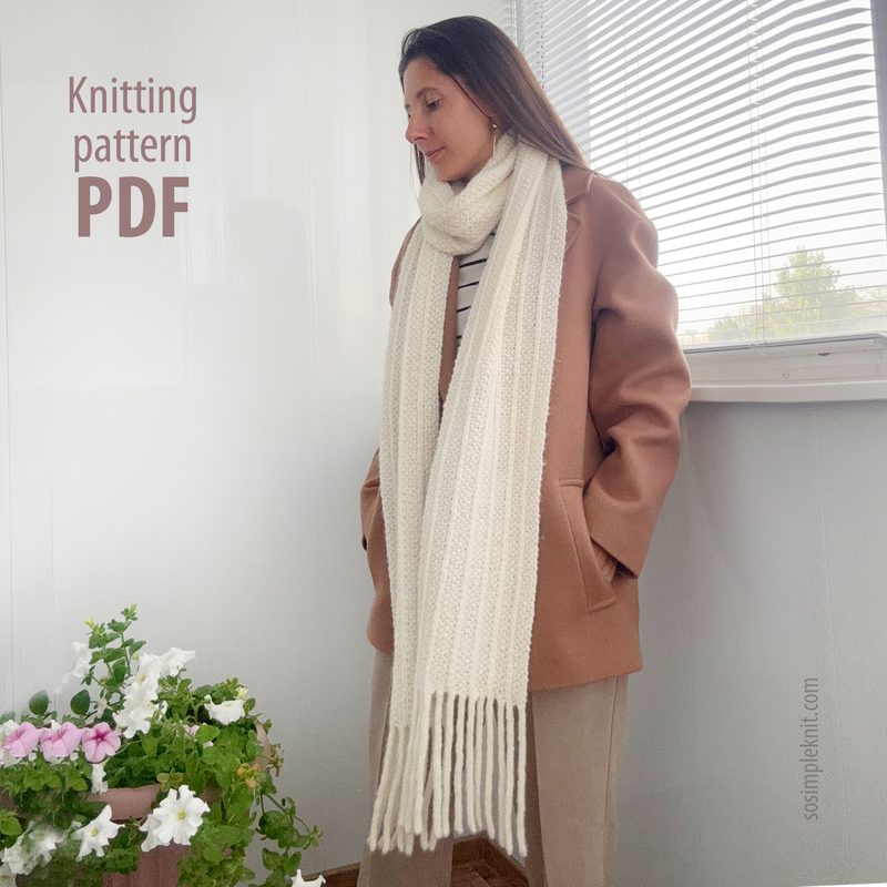 A woman stands against a light-colored wall, wearing a brown oversized coat and a beige sweater. She has long, brown hair and is modeling a long, cream-colored reversible scarf that drapes down to her waist, featuring fringed ends. The background includes a window partially covered by blinds, allowing soft natural light to enter, and there are pink and white flowers in a pot on the ledge. Text in the image indicates "Knitting pattern PDF."