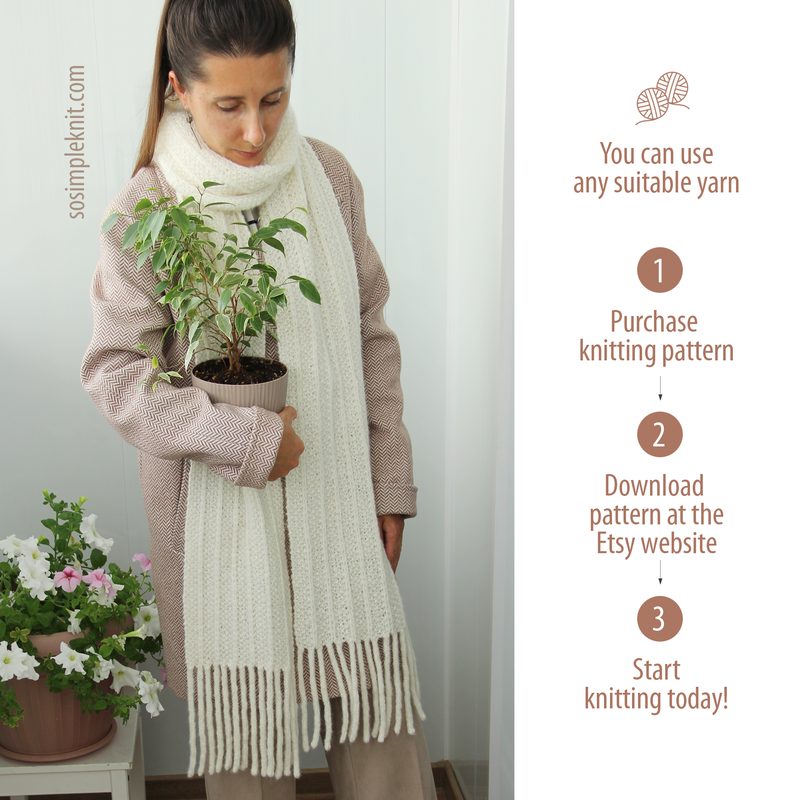 A woman stands indoors, wearing a light pink sweater over a white shirt, holding a potted plant in one arm. She is wrapped in a long, creamy white reversible scarf that features fringe at both ends. The background consists of white walls and indoor plants, including pink flowers at the bottom. The image has a soft and cozy aesthetic with natural lighting.
