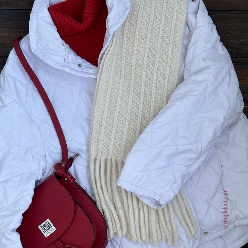 The image features a long, reversible knitted scarf in a cream color, draped over a white quilted jacket with a red collar. A red crossbody bag is positioned beside the jacket, complementing the scarf's color. The background consists of wooden planks, creating a warm contrast with the clothing items.