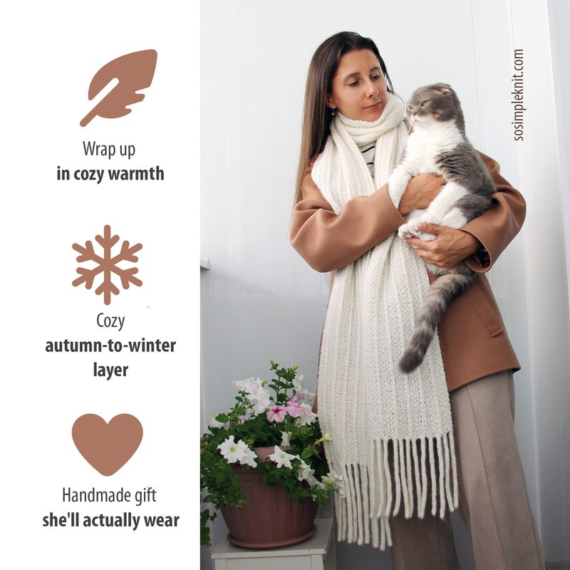 A woman in a beige coat stands indoors, holding a gray cat. She wears a long, knitted scarf that is cream on one side and a soft mauve on the other, with fringes at both ends. The background features a light-colored wall and a potted flowering plant with pink and white blooms on the floor. The image is framed to focus on the woman and her cat, with text elements on the left side describing the scarf's qualities.