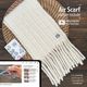 The image features a long, reversible knitted scarf displayed on a round woven mat. The scarf is light cream in color with a textured stitch pattern and has long, knotted fringes at one end. In the foreground, a tablet shows a partial view of a knitting video tutorial alongside two small cards, one of which includes the phrase "Air Scarf Knitting Pattern." The overall composition is warm and inviting, with natural textures and a cozy, hand-crafted feel.