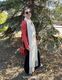 A woman stands outdoors beside a green pine tree, wearing a red leather jacket, black pants, and sunglasses. She is smiling and holding a red handbag. The long, reversible scarf she is wearing is white, featuring fringes at the ends. The background includes grass and a pathway, with sunlight filtering through the leaves.