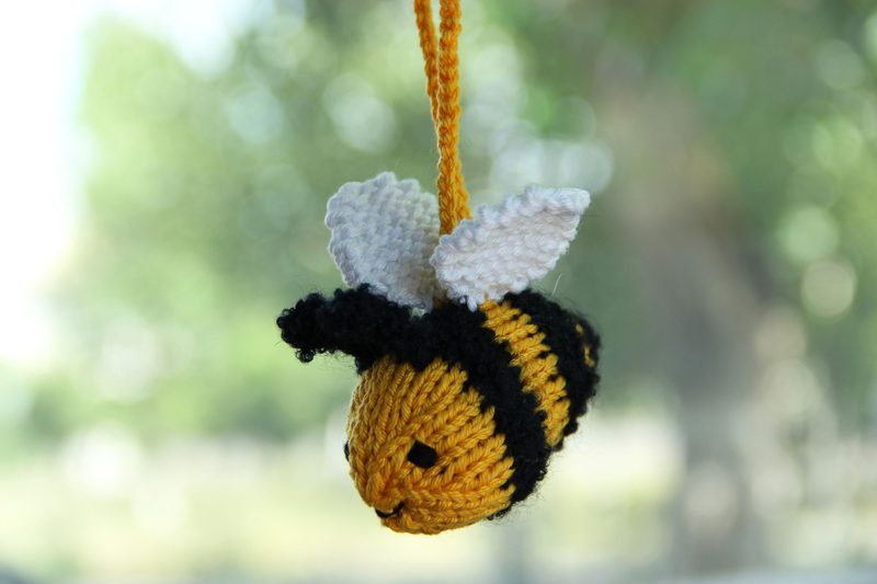 Knitted bee car hanging accessory made with needles, shown as a finished example of a knitting pattern; small yellow and black bee with white wings hanging on a string against a soft green blurred background