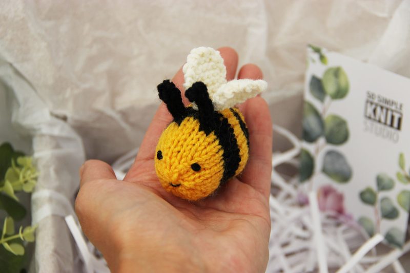 A hand holds a small, knitted honey bee, featuring a vibrant yellow body with black stripes and a white wing protruding from either side. The bee's face is simple, with two small black beaded eyes and a slight smile. The background includes soft, crinkled white tissue paper and a hint of greenery, creating a textured and natural setting. The image is bright and showcases the craftsmanship of the knitting pattern for beginners.