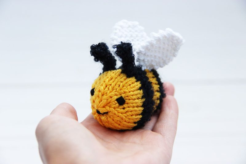 A small, knitted honey bee rests in a person's hand. The bee features vibrant yellow and black stripes on its body, with a round head and a smiling face. It has two small, black antennae and white, textured wings made of yarn. The background is softly blurred with a light color, emphasizing the bee's details. The overall style is cute and playful, suited for beginners in knitting.