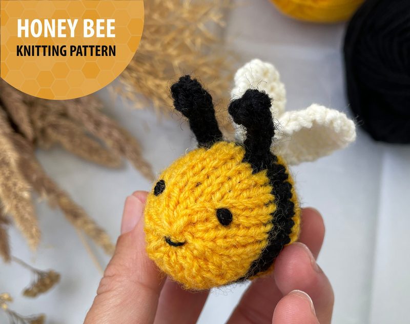 A close-up view of a small, round knitted honey bee, featuring a yellow body with black stripes, black antennae, and white wings. The bee has a simple smile stitched on its face. The background includes soft, neutral-colored elements, such as dried plants, and there are skeins of yarn in the background—one yellow and one black. In the top left corner, there is a circular overlay with the text "HONEY BEE KNITTING PATTERN." The overall composition is bright and cheerful, with a focus on the bee.