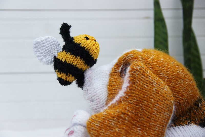 A close-up of a knitted honey bee resting on the nose of a knitted dog, showcasing the bee's yellow and black stripes, and white wings. The dog's fur is a mix of orange and white, with fine detailing in the knitting. The background features softly blurred green plants and a white wooden surface, adding a calm, natural ambiance to the scene.