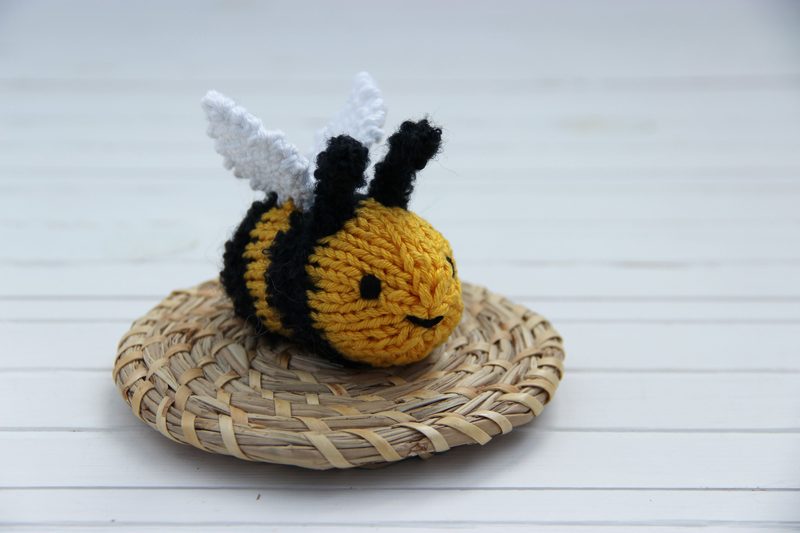 A small, handmade knitted bee lies on a round woven straw mat. The bee is primarily yellow with black stripes and features two white wings. It has a simple smiling face stitched onto its body. The background consists of light wooden planks, adding a natural and rustic aesthetic to the scene.
