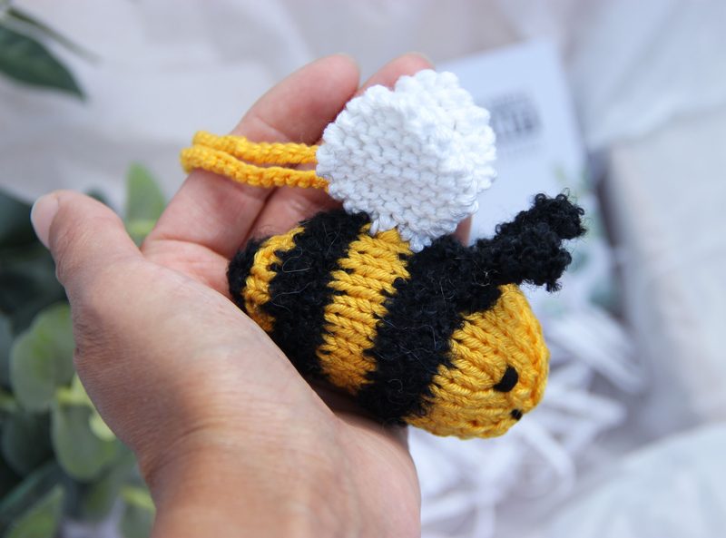 A knitted honey bee toy is being held in a person's hand, featuring a round body with yellow and black stripes, small black eyes, and a white, fluffy cloud-shaped wing. The background includes soft, neutral fabric and green leaves, providing a natural setting that highlights the bee's bright colors. The toy is framed closely, emphasizing its cute features and texture.