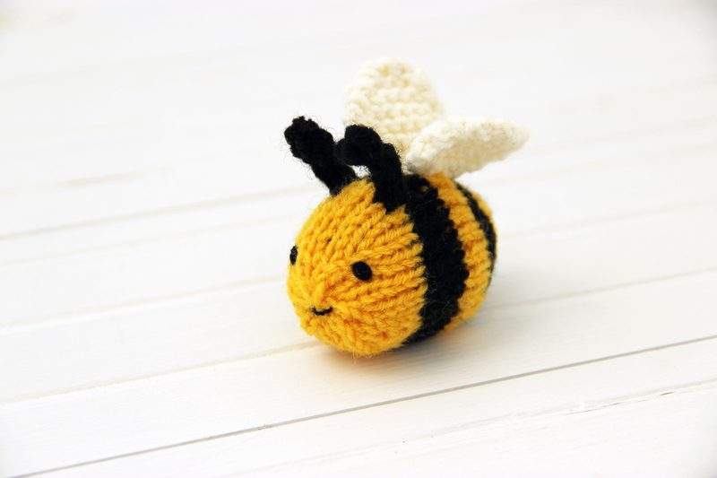 A knitted honey bee, featuring a round, yellow body with black stripes, small black eyes, and a stitched smile. It has two white wings made of knitted material attached to the top and is positioned on a white wooden surface, creating a clean, bright aesthetic. The overall look is cheerful and playful, suitable for a beginner's knitting pattern.