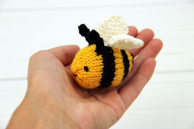 A small, knitted honey bee is held in a person's hand, featuring a rounded body with yellow and black stripes. The bee has a small smile and two black safety eyes. Two white wings made from yarn are attached to the top of the bee. The background is a light, neutral-colored wooden surface, emphasizing the bee's bright colors. The hand is positioned near the center of the image, with fingers gently cradling the bee.