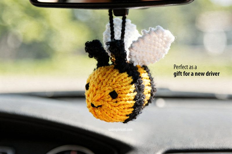 Knitted bee car mirror hanger made with needles, shown as a finished example of a knitting pattern; a small yellow and black bee with white wings hanging from a rearview mirror inside a car, with a softly blurred green view outside the windshield
