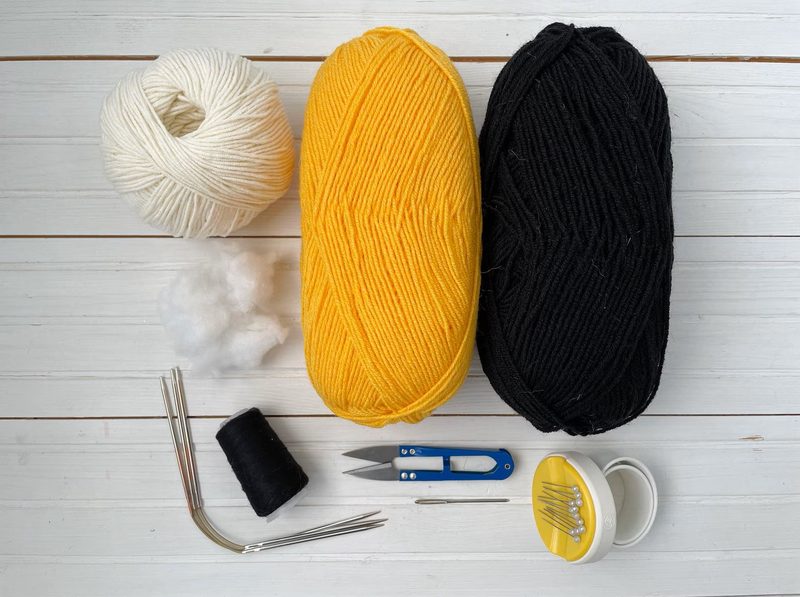 The image features two large skeins of yarn, one yellow and one black, placed side by side on a light wooden surface. A smaller white skein of yarn is positioned above them. Nearby, there is a small pile of white cotton stuffing, a black sewing thread spool, a craft knife, a metal needle, a set of knitting needles, and a small container with various sewing needles inside. The arrangement is organized, showcasing the materials needed for the honey bee knitting pattern.