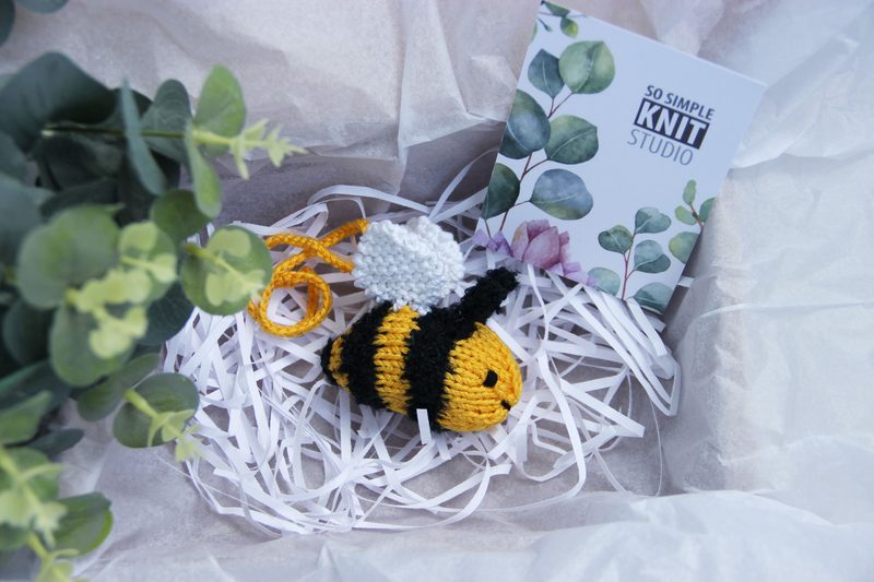 A knitted honey bee in yellow and black, with a white pom-pom for wings, is positioned among crumpled white paper shreds in a light brown box. Next to the bee is a small card featuring a botanical design with the text "So Simple Knit Studio" in a modern font. Surrounding the box are green leaves and greenery, creating a soft, natural framing for the image. The overall color palette includes earthy tones of yellow, black, green, and white.