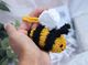 A knitted honey bee toy is being held in a person's hand, featuring a round body with yellow and black stripes, small black eyes, and a white, fluffy cloud-shaped wing. The background includes soft, neutral fabric and green leaves, providing a natural setting that highlights the bee's bright colors. The toy is framed closely, emphasizing its cute features and texture.