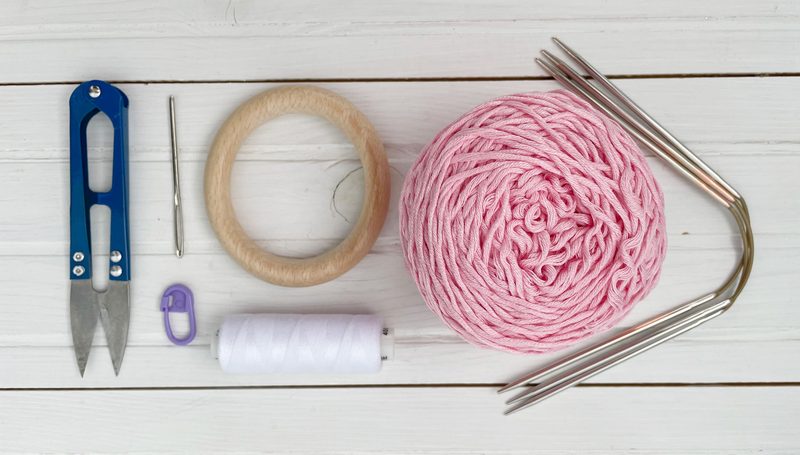 The image shows a flat lay arrangement of crafting materials for a knitting project. A circular wooden ring is positioned centrally, surrounded by a pink ball of yarn, a pair of blue scissors, a metal stitch marker, a white sewing thread spool, a small metal needle, and a set of silver knitting needles. The background features light-colored wooden planks. Each item is clearly visible and neatly organized, emphasizing the tools and materials needed for the Bunny Ears Teething Ring pattern.
