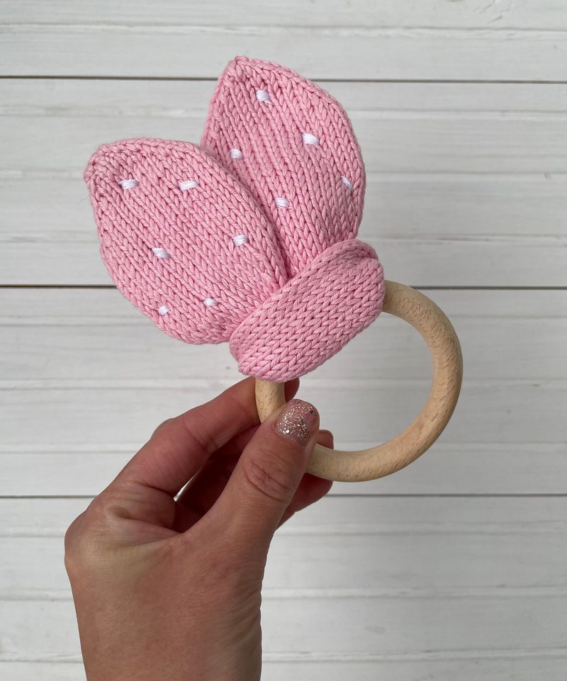 A hand displays a knitted teething ring featuring large, pink bunny ears with white dots and a textured base, attached to a smooth wooden ring. The background consists of light-colored wooden planks.