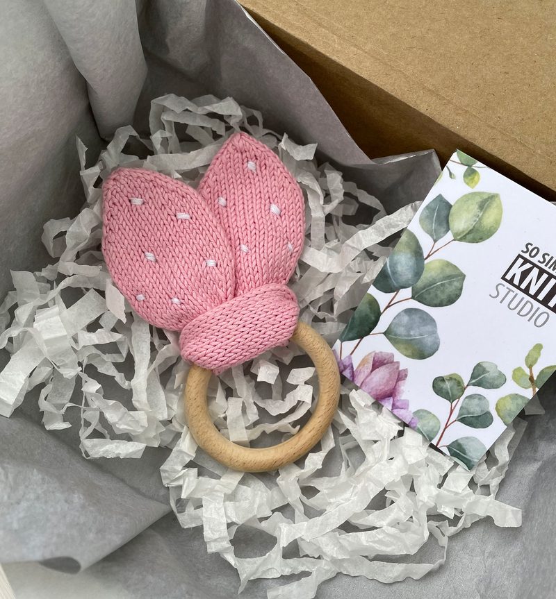 A soft pink knitted teething ring shaped like bunny ears rests on crumpled white tissue paper inside a cardboard box. The teething ring features white dot patterns on the ears and is attached to a natural wood ring. Next to the teething ring is a small card with a floral design and text that reads "Sosita Knit Studio." The overall setting is neutral and organized, highlighting the product's texture and color.