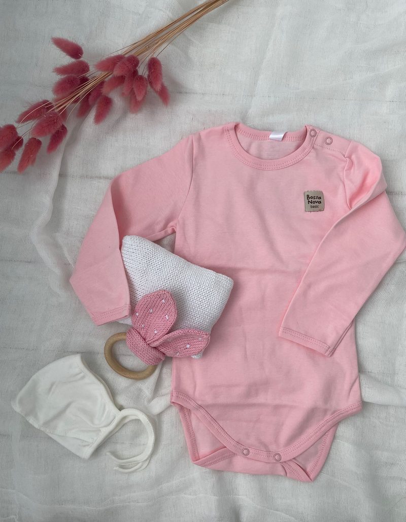 The image features a soft pink long-sleeve onesie with a small logo on the chest, a white textured bag with a bunny-ear shaped embellishment, a wooden teething ring, and a white headband. The background consists of a light, neutral fabric, and there are soft pink decorative accents, such as dried flowers, adding a gentle touch to the composition. The items are neatly arranged, highlighting their colors and textures.