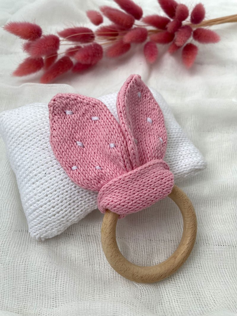 The image features a knitted teething ring with bunny ears in soft pink, resting on a white textured cushion. The teething ring has a circular wooden base and the ears are detailed with white stitching, giving a subtle polka dot appearance. In the background, there are dried pink flowers slightly out of focus, adding a decorative touch. The overall composition is light and gentle, with soft colors and a cozy aesthetic.