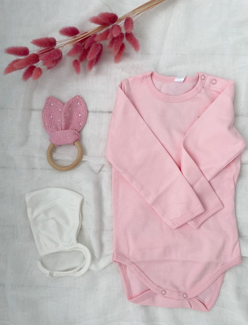 The image features a soft, light pink long-sleeve baby bodysuit positioned on a textured white fabric background. Next to the bodysuit, there is a white baby hat and a wooden teething ring adorned with pink bunny ears, embellished with small white dots. The composition includes a sprig of dried pink flowers, adding a delicate touch to the scene.