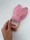 Hand holding a pink knitted bunny ears teething ring with a wooden ring, knitting pattern product image on a clean white background
