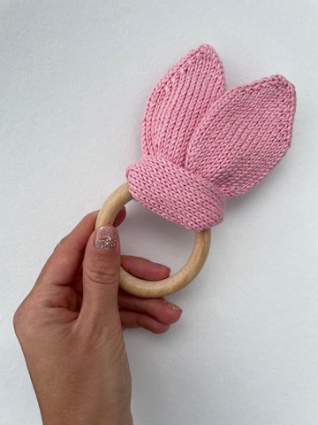 Hand holding a pink knitted bunny ears teething ring with a wooden ring, knitting pattern product image on a clean white background