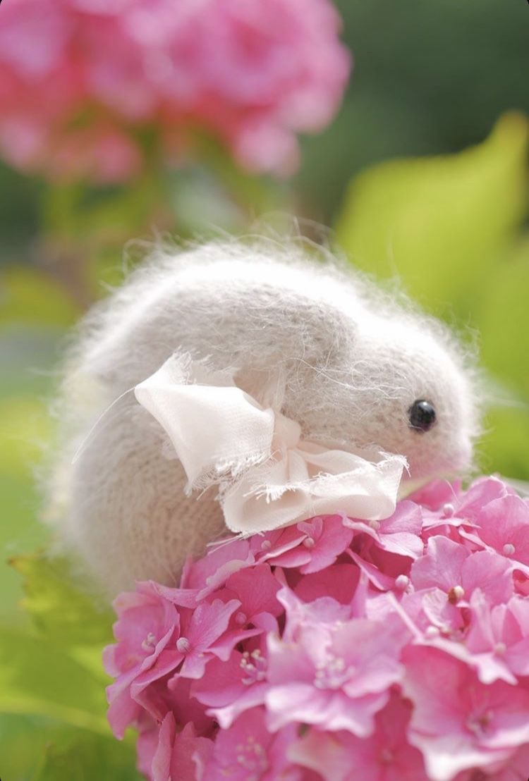A white, fluffy bunny toy with a pink ribbon sits among vibrant pink hydrangea flowers, showcasing soft details and a gentle expression. The background is blurred, featuring green leaves that frame the subject, creating a serene and colorful composition.