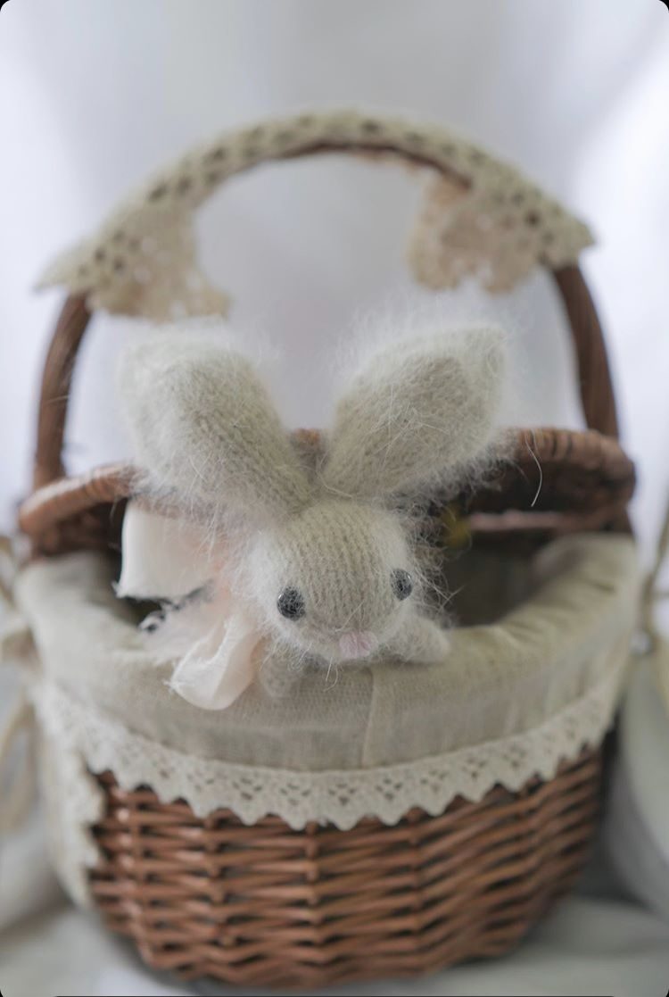 A small, hand-knitted bunny toy with a fuzzy texture sits in a woven wicker basket, which has a soft, cream-colored lining and lace trim. The bunny is light gray with large ears and a slightly pink nose, and it is adorned with a pale ribbon around its neck. The background is soft and blurred, enhancing the focus on the bunny and basket.