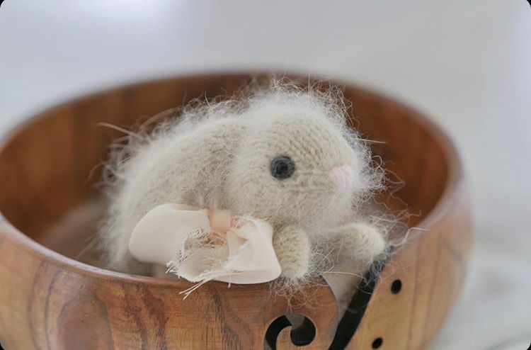 A small, handmade knitted bunny toy with a soft, fluffy texture is positioned inside a wooden bowl. The bunny is primarily a light beige color, featuring a pale pink nose and surrounded by a soft, pale pink bow tied around its neck. The background is blurred, emphasizing the bunny and the warm tones of the wooden bowl, which has a simple, curved design.
