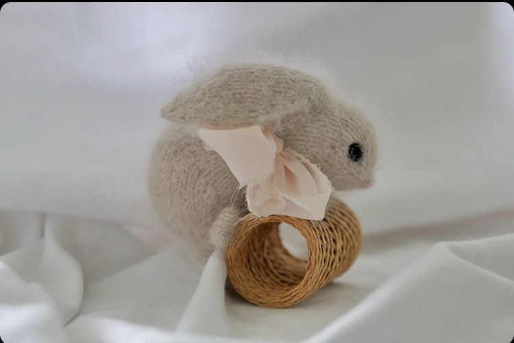 A soft, light beige knitted bunny with small black eyes and a pink satin bow sits beside a small woven basket. The bunny is turned slightly to one side, highlighting its rounded body and floppy ears. The background is soft white fabric, creating a gentle and calm atmosphere. The scene focuses on the bunny and the basket, with a shallow depth of field emphasizing the subjects.
