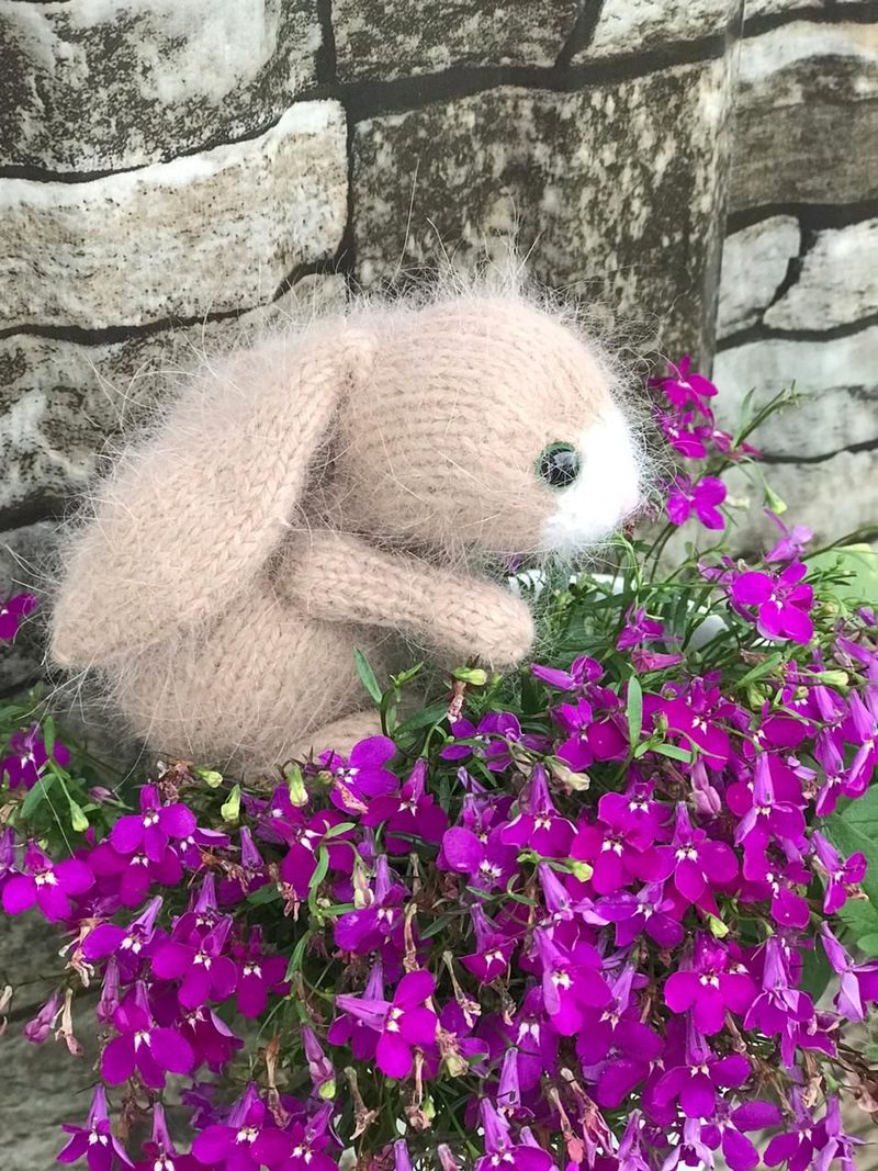 A small, knitted bunny toy in a soft beige color with prominent floppy ears and a white snout, sits nestled among vibrant purple flowers. The bunny's body has a fuzzy texture, and it is positioned to the side. The background features a stone wall with a rustic texture, contrasting with the colorful blooms. The overall composition focuses on the bunny against the floral setting.