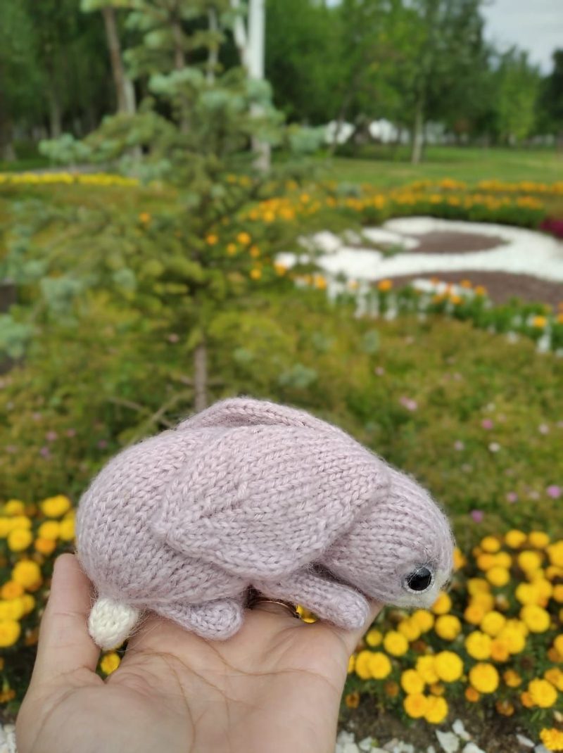 A small, knitted bunny toy in soft pink yarn is being held in a hand, set against a vibrant garden background with various flowers and greenery. The bunny features small black eyes and a white tail, with its ears slightly folded backward. The image is framed to highlight the toy in the foreground, while the colorful garden, filled with yellow marigolds and green plants, softly blurs in the background.