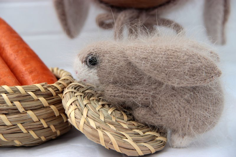 A fluffy, light brown knitted bunny toy sits next to a woven basket containing three orange carrots. The bunny has large black eyes and pink accents on its nose and ears. The scene is softly lit, highlighting the texture of the yarn. The background features a blurred out basket or other objects, keeping focus on the bunny and carrots in the foreground.