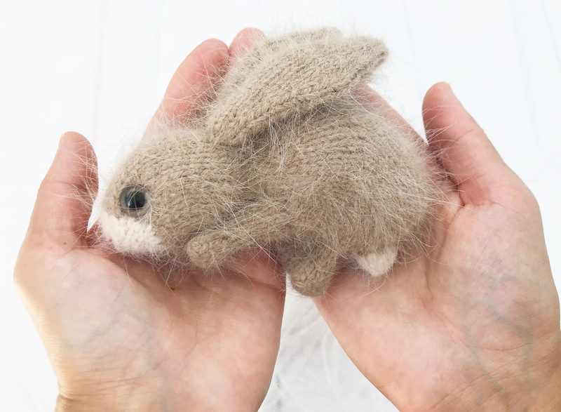 A small, handmade bunny toy is nestled in two hands, showcasing its soft, fuzzy texture. The bunny is light brown with darker brown accents on its ears and a white face. Its ears are upright, and it has large, black safety eyes. The hands are gently cupping the toy, contrasting against a light, neutral background. The overall composition emphasizes the toy's size and detail.