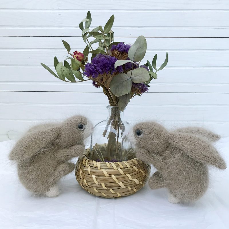 Two soft, beige bunny toys with round bodies and long ears sit on either side of a small woven basket containing dried flowers. The bunnies have a fuzzy texture, and their feet are white. The background features a simple white wooden wall, creating a light and airy atmosphere. The arrangement is symmetrical, with the bunnies focused on the basket in the center, emphasizing a cozy, serene setting.