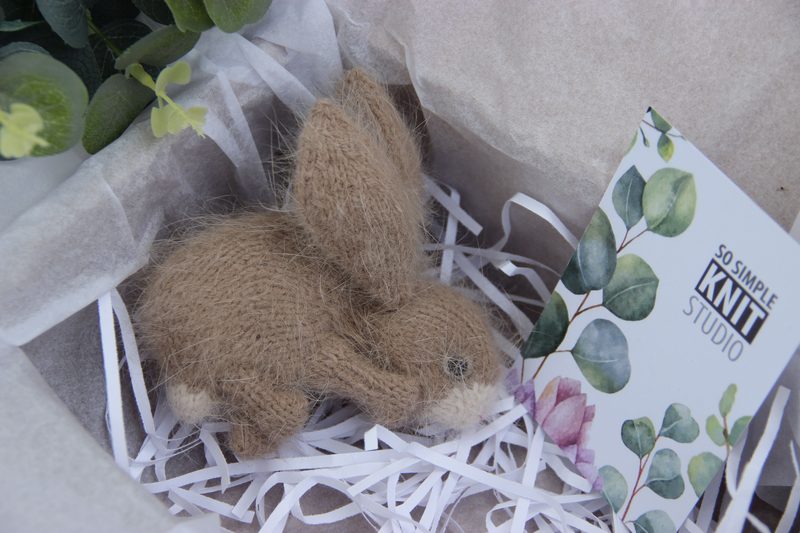 A small, knitted bunny toy made from brown yarn rests on a bed of white shredded paper, surrounded by light-colored tissue paper. The bunny features stitched facial details and upright ears. Beside it is a business card from "So Simple Knit Studio," adorned with green leaves and a floral design. The overall presentation is neatly arranged in a shallow, pale box, with some greenery visible in the background.