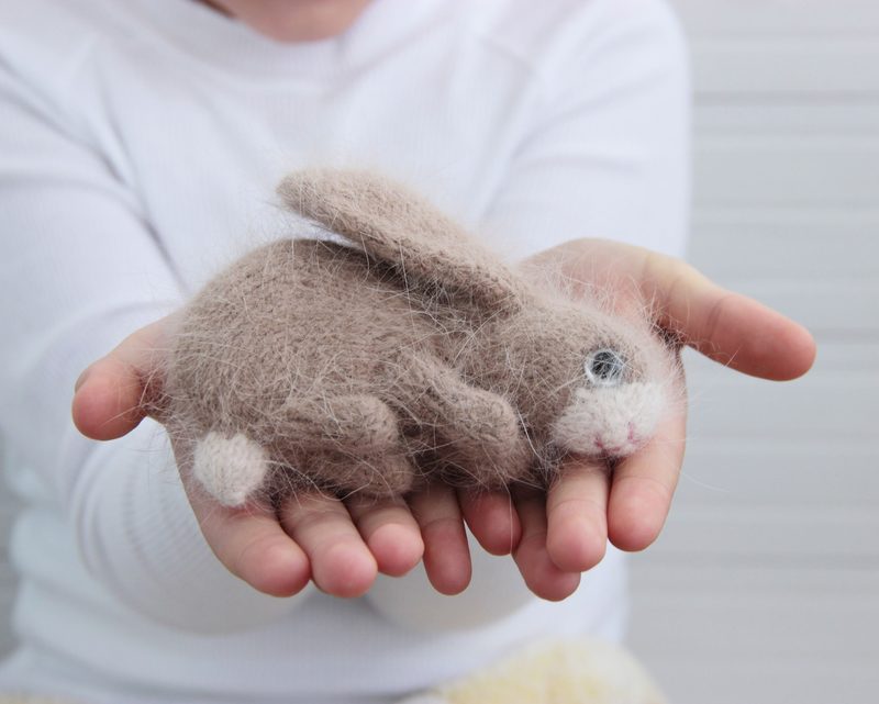 A person’s hands gently cradle a small, handcrafted bunny toy made from soft, felted fibers. The bunny has a light brown body, with a white underbelly, paws, and a small pink nose. Its eyes are depicted with small black beads, giving it a cute expression. The background is a subtle, light-colored wood texture, which emphasizes the bunny's soft colors and details. The person's fingers are delicately placed around the toy, showcasing its size and softness.
