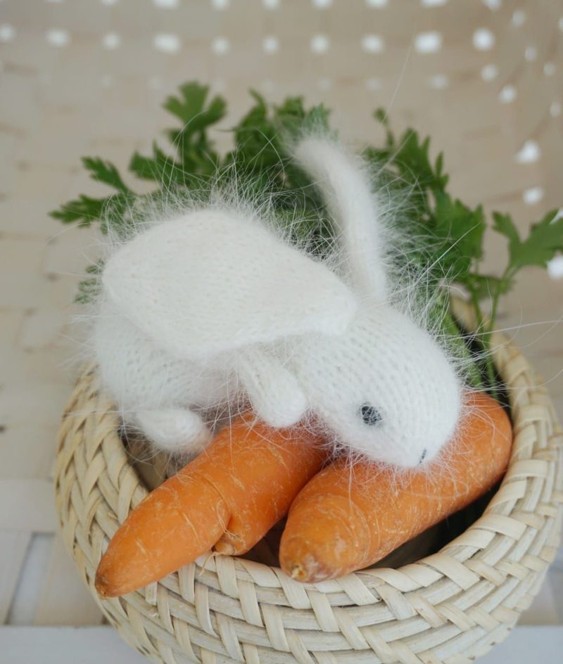 A small, white knitted bunny with long ears sits atop two orange carrots, nestled in a woven basket. The bunny features a fluffy texture and has a small pink nose, while green parsley leaves are visible alongside the carrots, adding a touch of greenery. The background is softly blurred, focusing attention on the bunny and vegetables.
