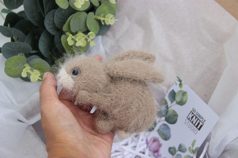 A small, handmade brown bunny toy with soft, fuzzy texture is held in a person's hand. The bunny features large, expressive eyes and tiny pink details on its nose and ears. It has small, rounded ears and fluffy wings. In the background, there are green leaves and a white tissue paper, along with a card that reads “Go Simple Knit Studio.” The overall scene is softly lit and creates a cozy, inviting atmosphere.