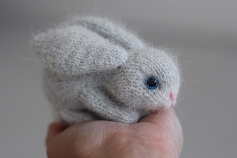 A small, grey knitted bunny toy with prominent ears and blue plastic eyes is cradled in a person's hand. The bunny has a soft, fuzzy texture, with a small pink nose and a compact, round body. The background is blurred and light-colored, emphasizing the bunny in the foreground. The hand is partially visible, showcasing the toy's size and adding a sense of warmth to the image.