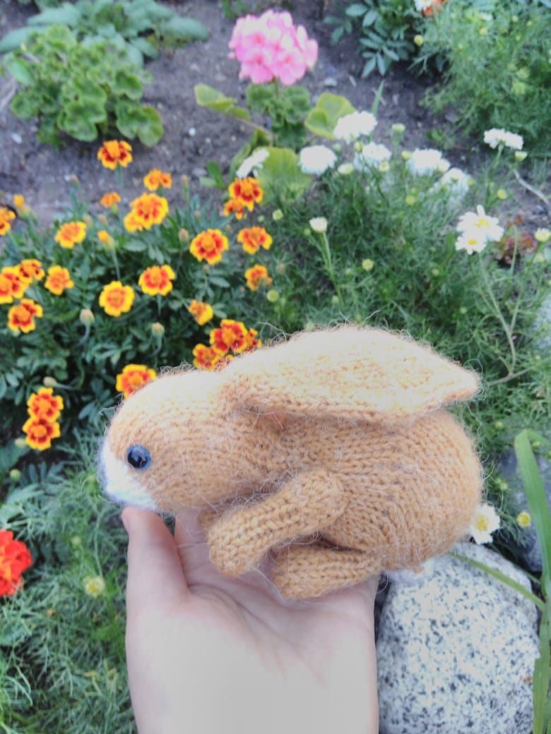 The image shows a knitted bunny toy held in a hand, featuring a soft brown body with a white muzzle and large ears. The toy is set against a vibrant garden background filled with multicolored flowers, including marigolds and pink geraniums, surrounded by lush green foliage. The hand slightly lifts the bunny, while the flowers create a lively and colorful setting around it.